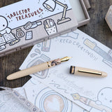Tabletop treasures box with a pen, ink bottle, and drawings on a wooden surface.